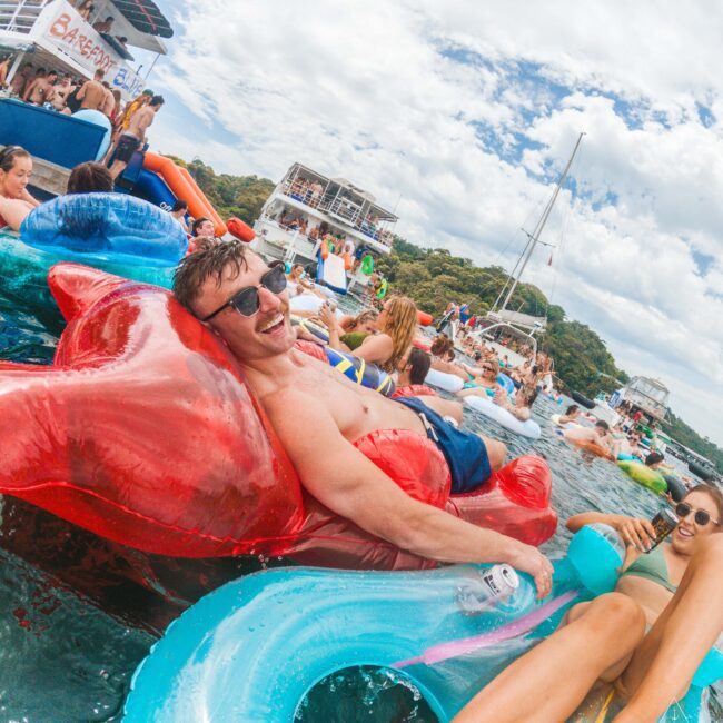 A man in sunglasses lounges on a red inflatable in the water, smiling at the camera. Around him are other people on colorful floaties, boats, and a party atmosphere under a partly cloudy sky.