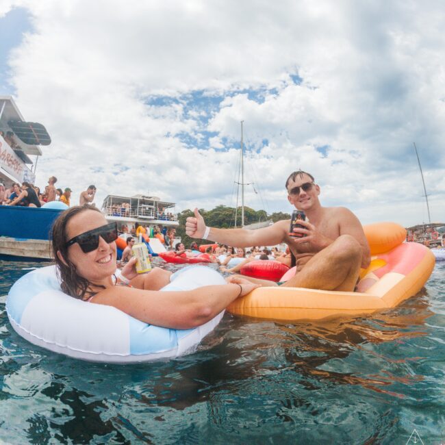 Two people in sunglasses relax on colorful pool floats in the water, smiling and giving thumbs up while holding drinks. Other floats and people, boats, and a partly cloudy sky are visible in the background.