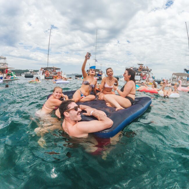 A group of five people in swimsuits laugh and pose for a selfie while relaxing on an inflatable float in a lake, surrounded by boats and others enjoying the water under a partly cloudy sky.