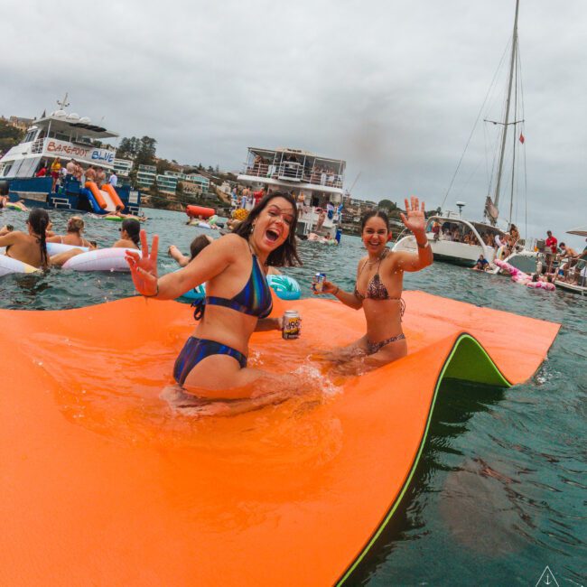 Two women in swimsuits smile and wave while sitting on a floating mat in the water. Boats and other people on inflatables are visible in the background under a cloudy sky.