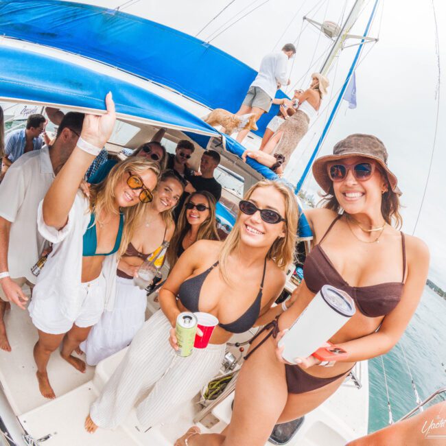 A group of smiling young adults in swimsuits and sunglasses enjoy a lively boat party on the water, holding drinks and posing for the camera under a blue canopy on a sunny day.
