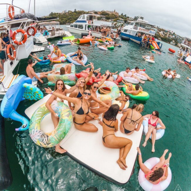 A group of people in swimsuits relax on colorful pool floats in the water between boats, enjoying a lively party on a sunny day. Several boats and more people are visible in the background.