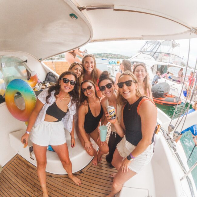 A group of seven smiling women in summer outfits pose together on a boat, with water, another boat, and other people in the background. A colorful inflatable ring hangs on the side, and several women are holding drinks.
