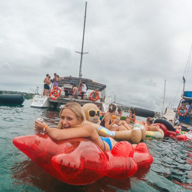 A woman in a swimsuit smiles while lounging on a red inflatable float shaped like lips in the water, with other people and floats nearby. A catamaran and cloudy sky are in the background.