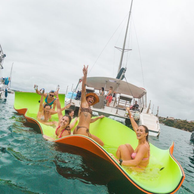 A group of smiling people in swimsuits relax on bright floating mats in the water, posing and waving near a sailboat on a cloudy day.