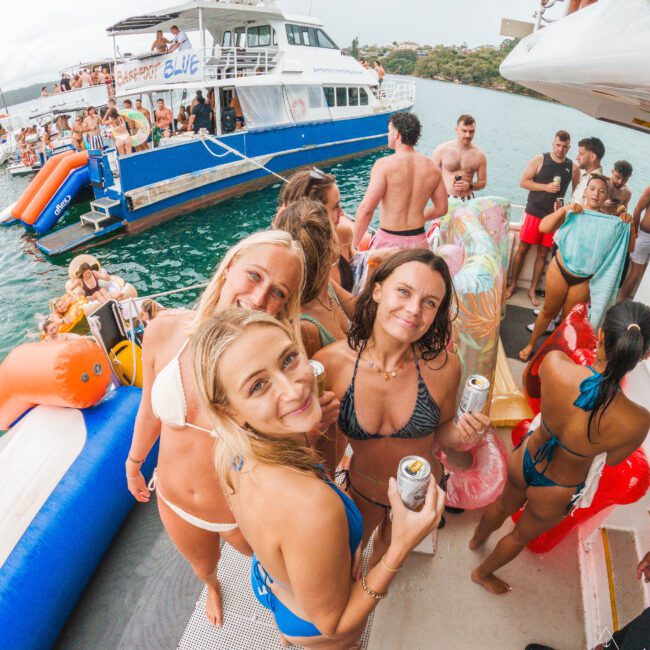 Three women in swimsuits smile and hold drinks on a crowded boat party, surrounded by people, pool floats, and another boat on the water in the background. The atmosphere is lively and festive.