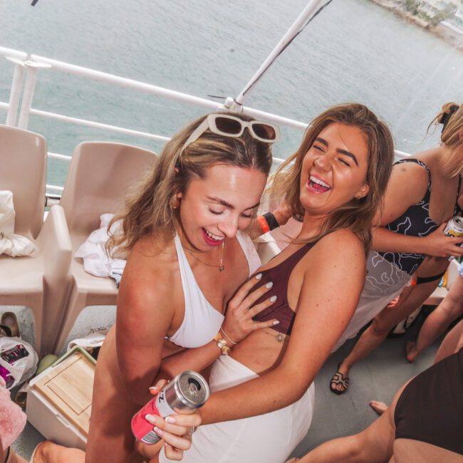 Two women in swimsuits laugh and pose together on a boat, holding drinks with a body of water and a boat in the background. People and bags are visible around them, creating a lively and joyful atmosphere.