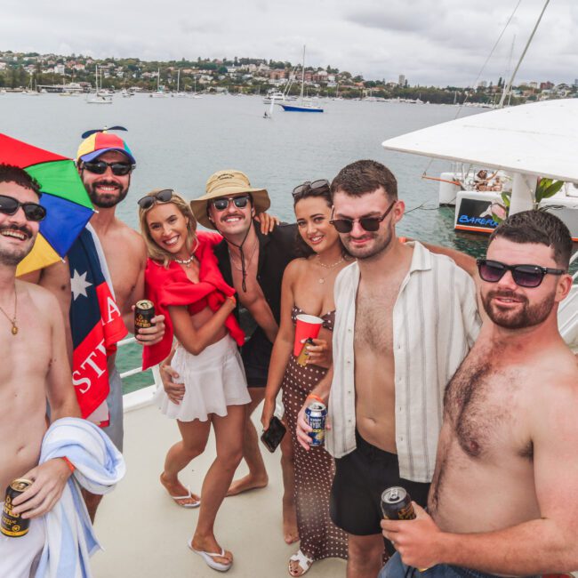 A group of seven friends, wearing swimsuits and summer clothing, smile and pose together on a boat. They hold drinks and stand by the railing, with water, boats, and a shoreline visible in the background.