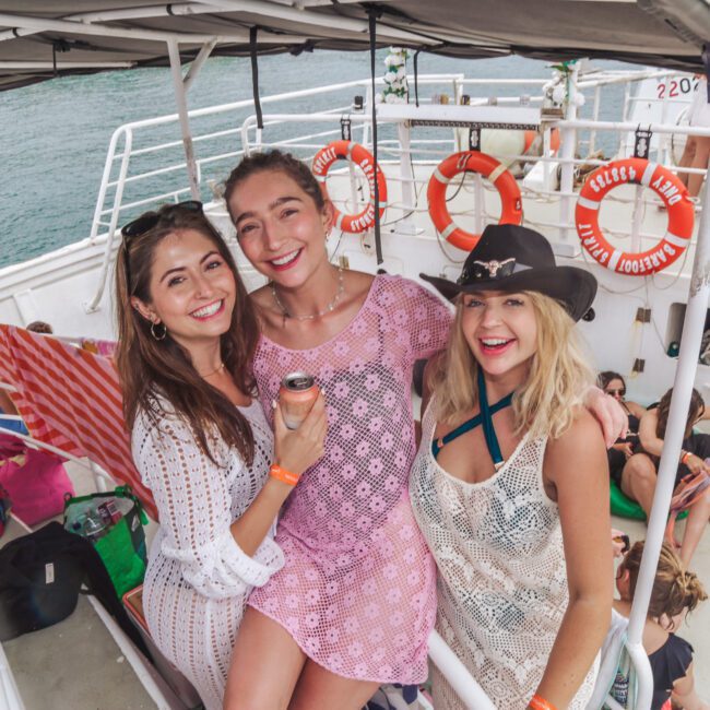 Three women smiling and posing together on a boat deck, wearing beach cover-ups and summer hats, with water and life rings visible in the background. Other people relax on the deck behind them.