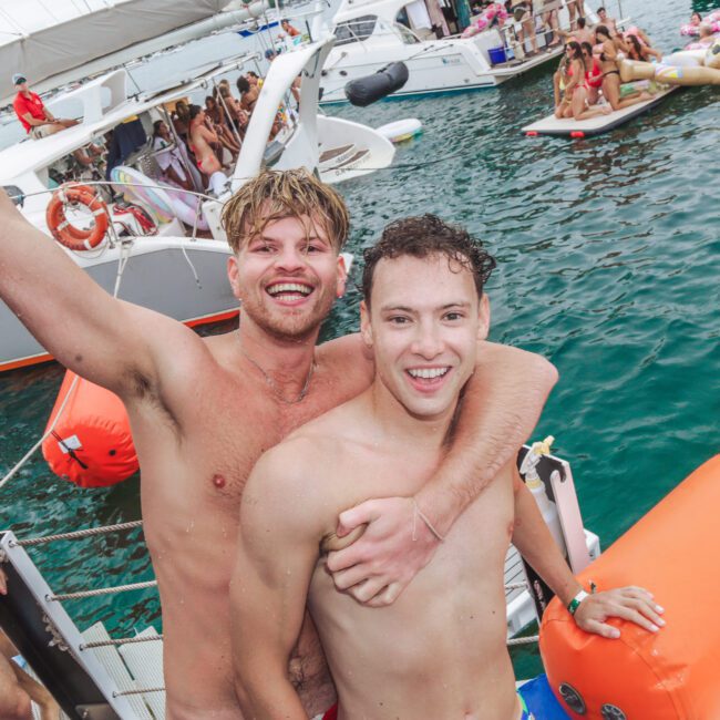 Two smiling shirtless men pose happily on the edge of a boat during a lively party. Other people are swimming, lounging on inflatables, and relaxing on nearby boats in the background. The atmosphere is festive and fun.