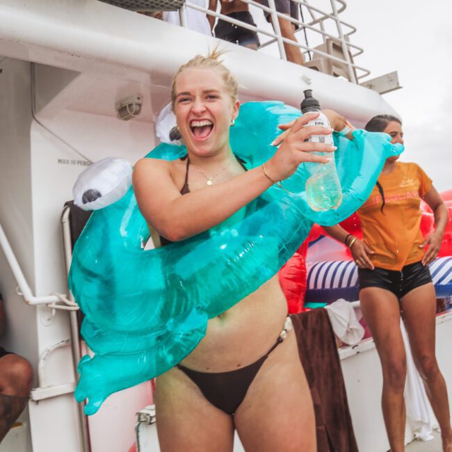 A smiling woman in a black bikini holds a drink and wears a blue inflatable pool float on a boat, surrounded by other people enjoying a party.