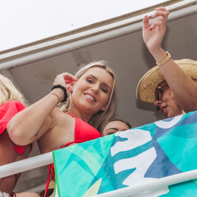 Three women stand on an outdoor deck behind a white railing. One smiles at the camera in a red bikini top, another wears a straw hat and sunglasses, and a third looks away. A colorful blue-green towel hangs on the railing.