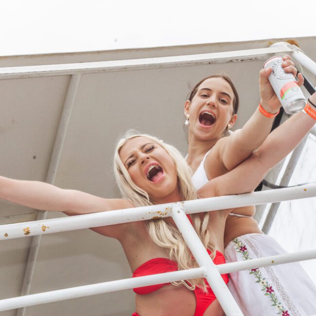 Two young women smiling and holding drinks, standing behind white railings outdoors. One wears a red bikini, the other a white outfit. Both have arms raised and look happy and excited.