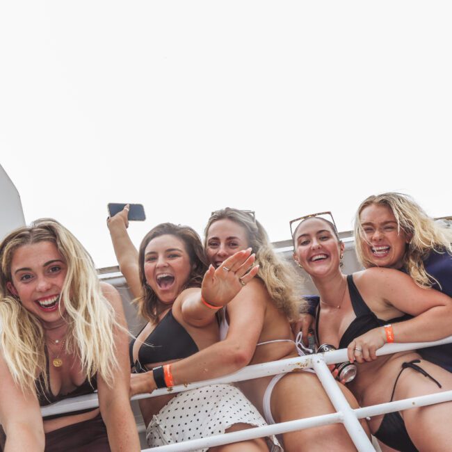 Five smiling young women in swimsuits and casual wear lean over a white railing, waving and posing for the camera. They appear joyful and energetic, with overexposed sky in the background.