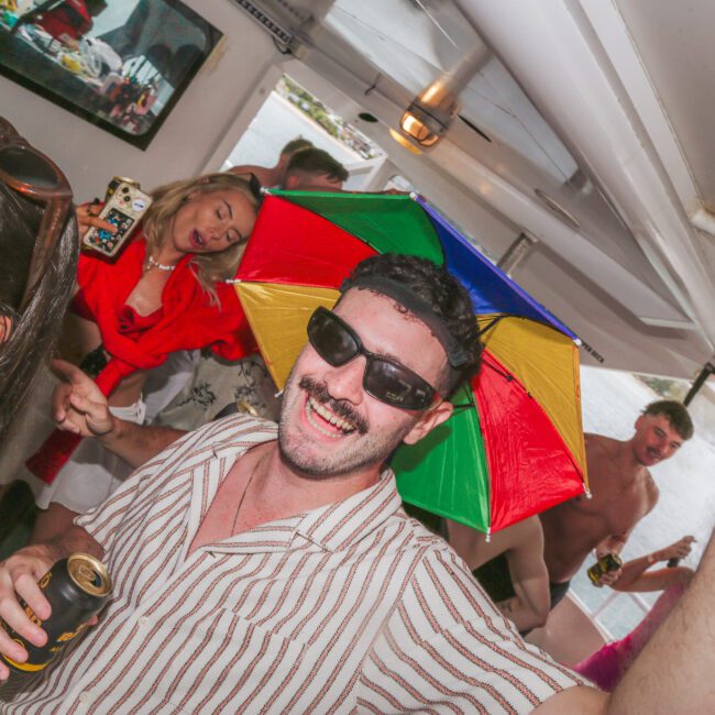 A man wearing sunglasses and a colorful umbrella hat smiles and raises his hand at a lively indoor party with other people around him, some holding drinks.