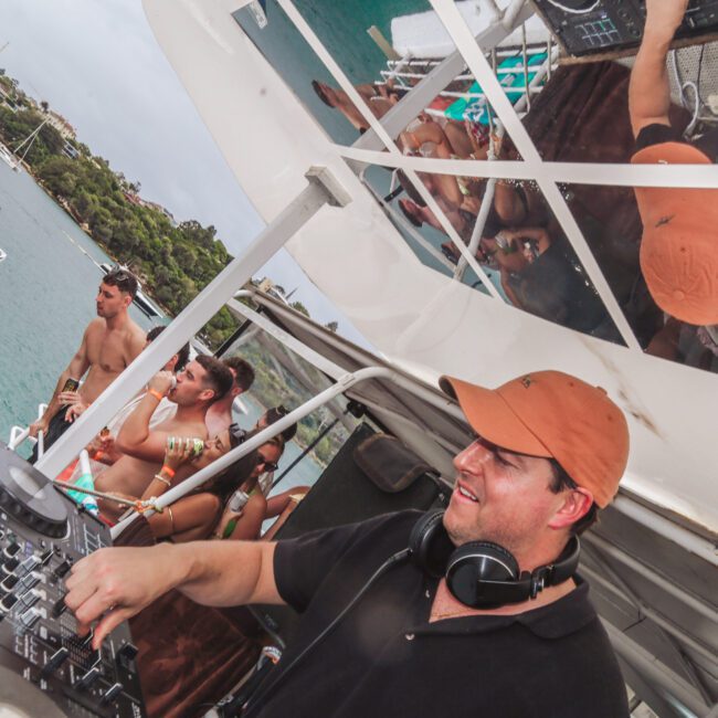 A DJ in a brown cap and black shirt plays music on a boat, with headphones around his neck and people dancing in swimsuits behind him. The scene is reflected in the boat's ceiling. Water and another boat are visible nearby.