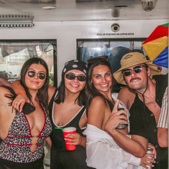 Six young adults wearing sunglasses and summer clothing smile and pose together at a party on a boat; one person holds a rainbow umbrella hat, and another holds a red cup.