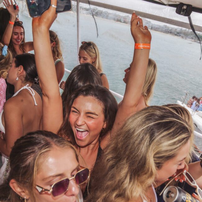A group of young women are having fun and dancing on a crowded boat, holding drinks and raising their arms while smiling and laughing. The boat is on the water with other boats visible in the background.