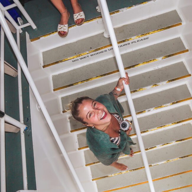 A woman smiles up at the camera while standing barefoot on a white staircase. She is wearing a green top and has a camera around her neck. Another person’s feet are visible at the top of the stairs.