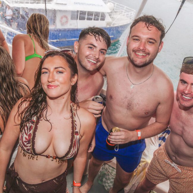 A group of four smiling young adults in swimwear pose together on a boat, with other people and a large boat visible in the background on the water. The scene looks lively and festive.