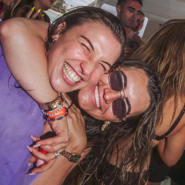 Two smiling women hug and pose for a photo at a crowded, lively indoor party. Both wear swimwear and festival wristbands. Others in the background are laughing and having fun.