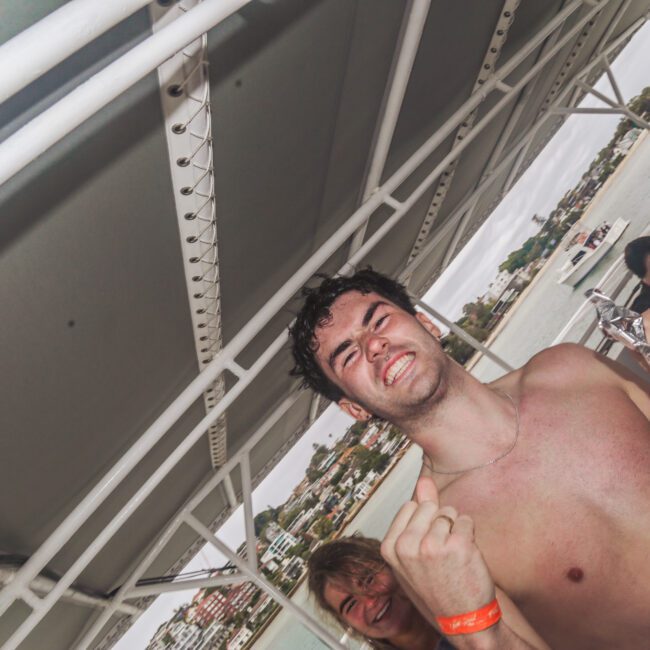 A young man with wet hair, wearing a red wristband and holding a foil bag, stands shirtless on a boat, smiling enthusiastically. People and water are visible in the background under a cloudy sky.