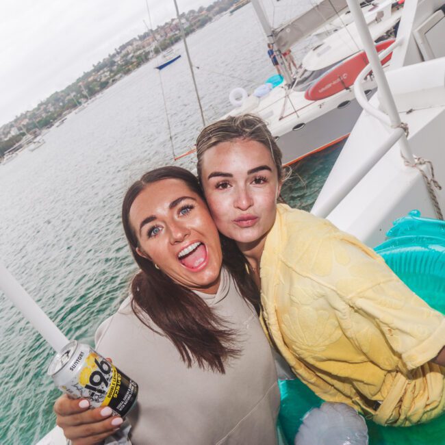 Two young women smile and pose on a boat. One wears a beige top and holds a can, while the other wears yellow and sits on a blue pool float. Water and other boats are visible in the background.
