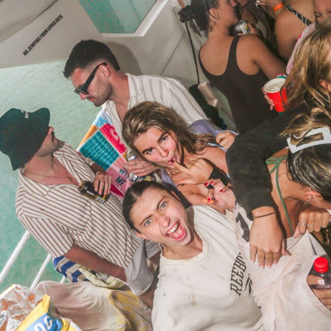 A group of young people smiling and having fun at a lively boat party, holding drinks and snacks, with water visible in the background.