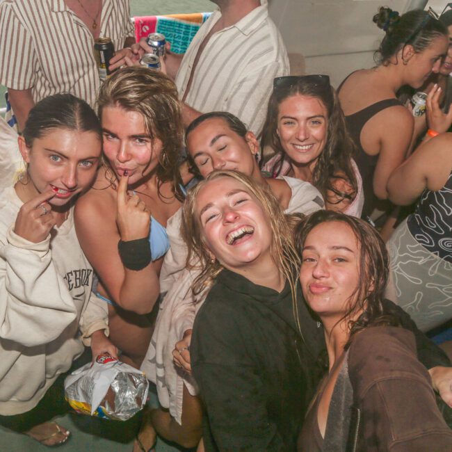 A group of young women in swimsuits and casual clothes smile and pose together on a boat, some holding drinks and snacks, surrounded by other people enjoying a lively atmosphere.
