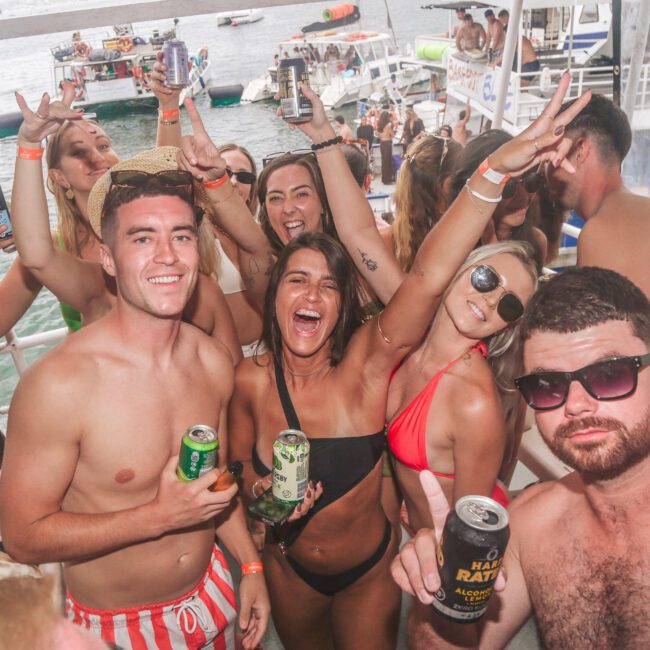 A group of young adults in swimsuits smiling and holding drinks on a boat, enjoying a party. Some make hand gestures and pose for the camera. There are boats and water in the background.