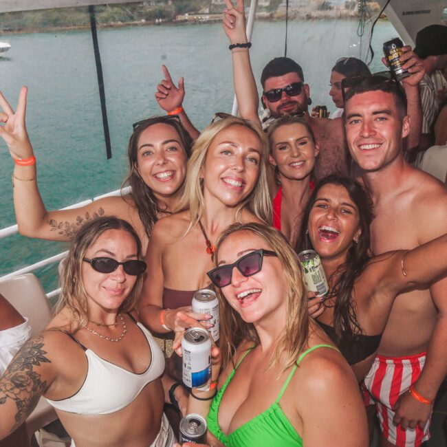 A group of young adults in swimsuits smile and pose for a photo on a boat party, holding drinks with the ocean and another boat visible in the background. The atmosphere is lively and joyful.