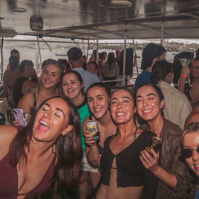 A group of young women smile and pose for a selfie while holding drinks at a crowded boat party. Other people are in the background, and water and buildings are visible through the windows.