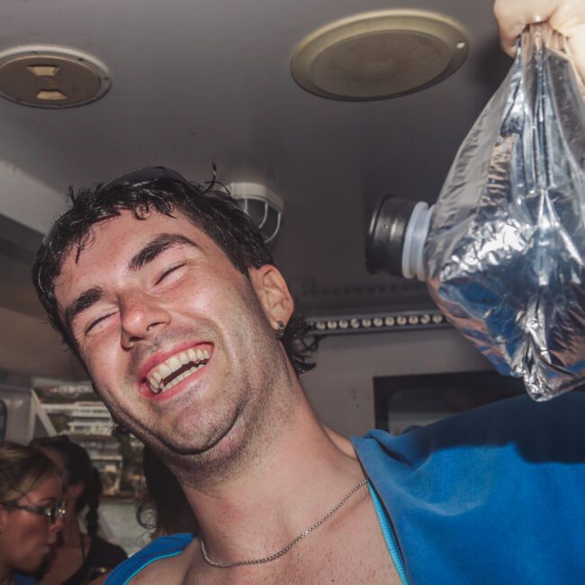 A smiling man with wet hair holds up a plastic bag filled with liquid and a bottle top, standing on a boat with people and buildings visible in the background.
