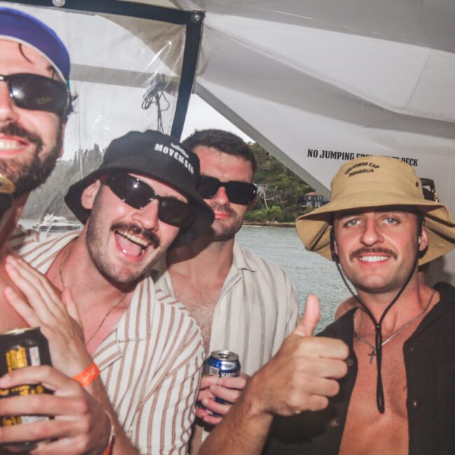 Four men in casual clothing and hats pose for a cheerful photo on a boat, smiling and holding drinks. One gives a thumbs up. Water and greenery are visible outside the window in the background.
