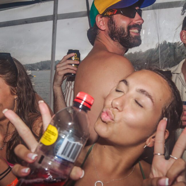 A group of young adults at a lively outdoor party; a woman in the foreground poses with a peace sign and kissing face, while others smile and hold drinks. The setting appears to be a boat or near water.