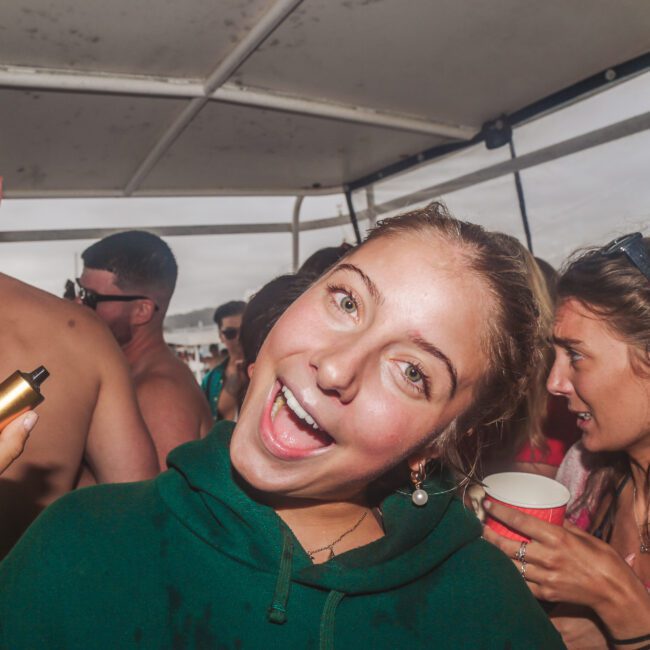 A smiling young woman in a green hoodie poses with a vape on a crowded boat, surrounded by people enjoying a party; some hold drinks, and the background shows water and a cloudy sky.