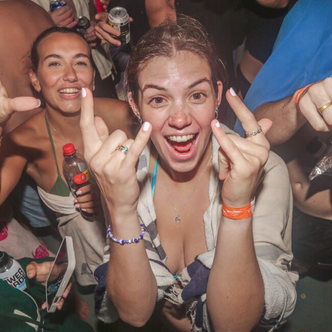 A group of smiling people at a lively party, with a woman in the center making playful hand gestures toward the camera. People hold drinks and wear swimsuits and wristbands, suggesting a fun outdoor or pool event.