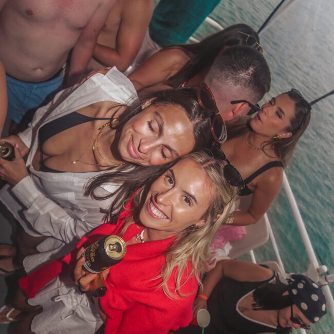 A group of young adults enjoy a boat party near the water. Two women in the foreground, one in red and one in white, smile at the camera holding drinks. Others mingle and relax in swimwear in the background.