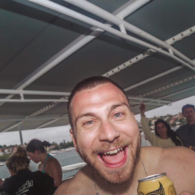 A smiling, shirtless man holding a can poses in the foreground on a boat with people and water visible in the background under a covered deck. The mood is lively and festive.