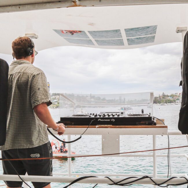 A DJ wearing headphones stands behind a mixer on a boat deck, playing music. Large speakers flank him, and water and a cloudy sky with distant buildings are visible in the background.
