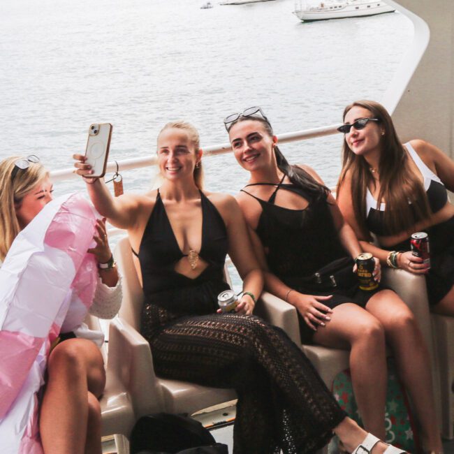 Four women sit on a boat, smiling and posing for a selfie. One holds a phone, another holds a pride flag. The sea and yachts are visible in the background. They appear happy and relaxed, wearing swimsuits and sunglasses.