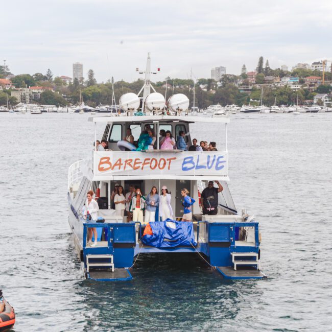 A white boat named "Barefoot Blue" is on the water with people standing on its deck, some waving. Smaller boats and numerous buildings are visible in the background under a cloudy sky.