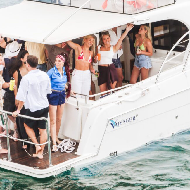 A group of people on a white yacht named "Voyager" are smiling and raising their drinks, enjoying a sunny day on the water. Some are dressed in swimwear, and the boat is surrounded by calm, blue-green water.