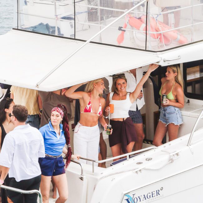 A group of people in swimwear and summer outfits enjoy drinks and socialize on the deck of a white boat named "Voyager" on the water. Some are smiling and laughing, creating a lively, festive atmosphere.