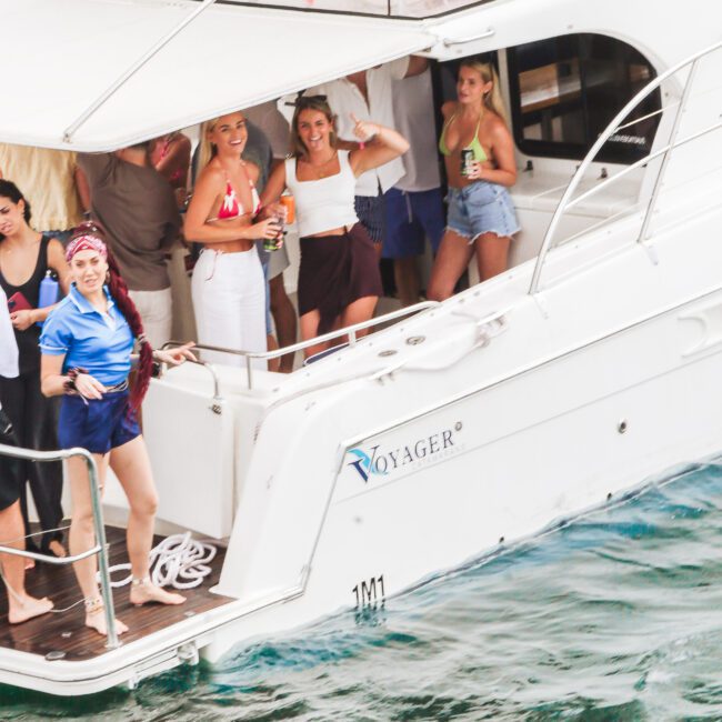 A group of people stand and smile on the deck of a white yacht named "Voyager" docked on clear blue water, holding drinks and wearing casual summer outfits.