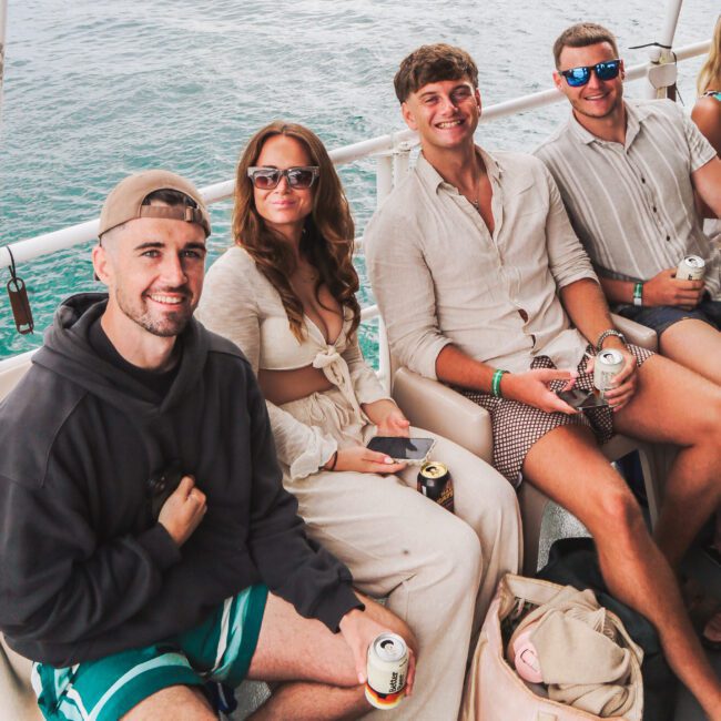 Four people sit closely together on a boat, smiling and holding canned drinks. The background shows calm water and boats. They are dressed in casual summer clothes and sunglasses. A beige tote bag sits on the floor near their feet.
