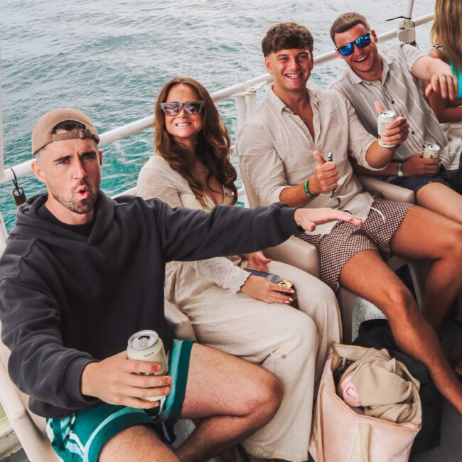 Four people sit and smile on a boat, holding canned drinks. They wear casual summer clothes and sunglasses, with water and boats visible in the background. One person gestures animatedly towards the camera.