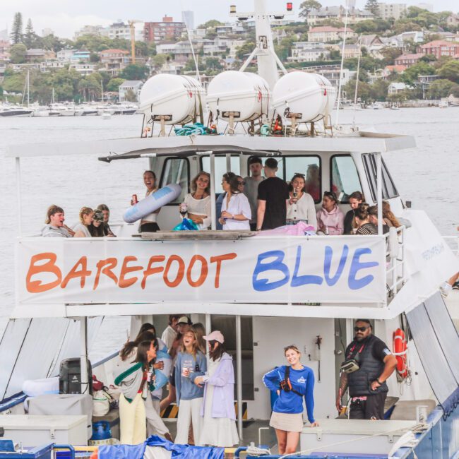 A group of people stands and socializes on a two-level boat named "Barefoot Blue," docked in a harbor with water, trees, and city buildings in the background. Some are looking at the camera and smiling.
