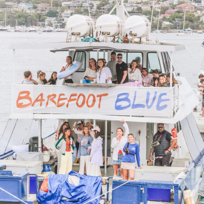 A group of people stand and socialize on the deck of a boat named "Barefoot Blue," docked by the water, with a residential area and trees visible in the background.