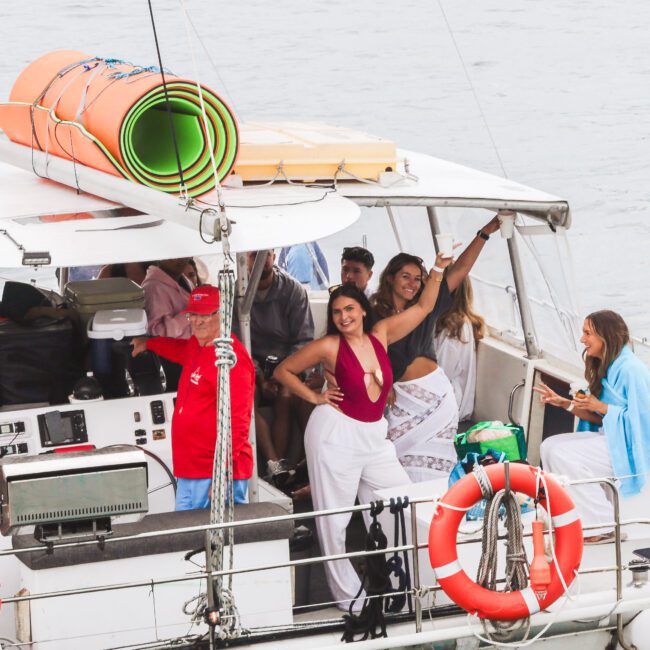 A group of people stand and pose on a white boat docked on the water. Some are smiling and wearing casual summer clothes. Colorful foam mats are rolled up on the roof, and a lifebuoy is attached to the railing.
